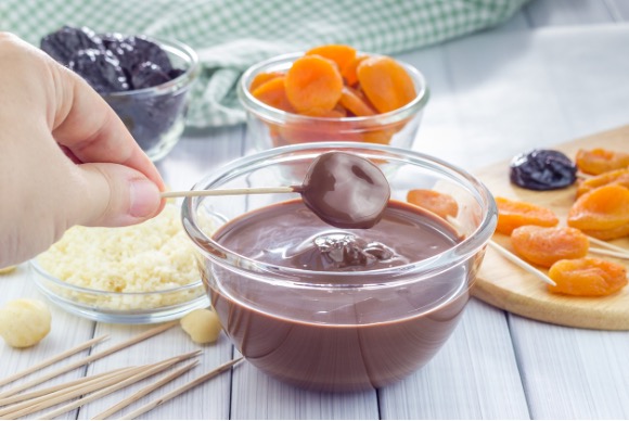 A person dipping a stick with fruit in a bowl of chocolate